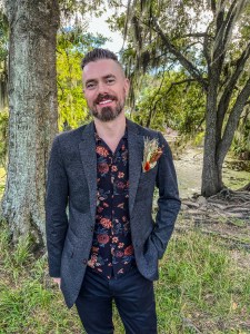 Photo of author Alexander Schneider. The author is standing in a clearing among trees, wearing a casual three-piece suit with a boutonnière.